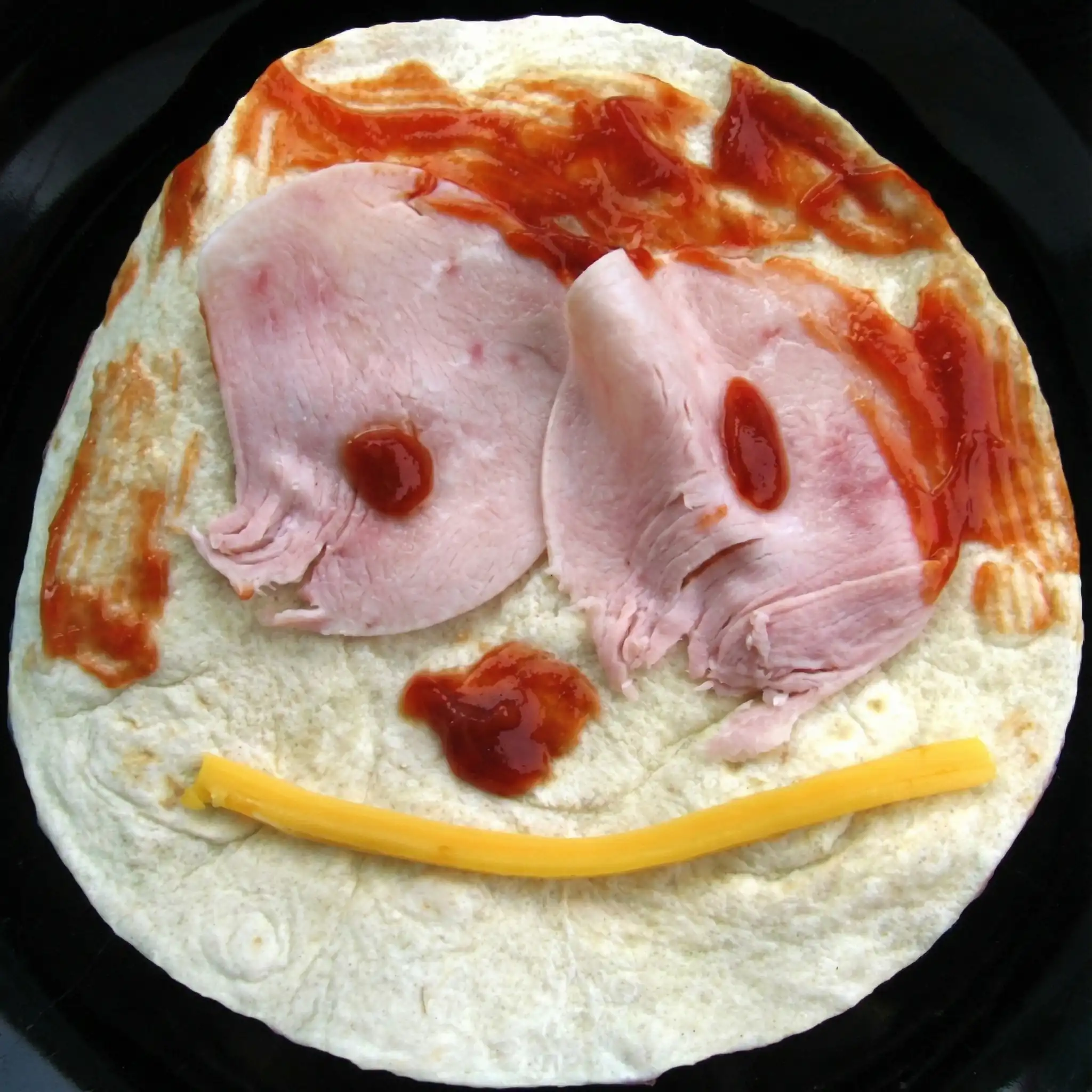 A round tortilla on a black background decorated as a face: two ham slices with ketchup dots as eyes, ketchup smeared as hair and nose, and a curved yellow cheese stick forming a smile.